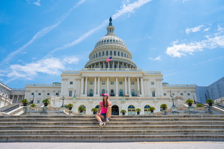a woman sits at the steps of the capitol building with the front of the building in the background