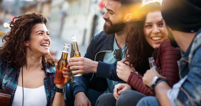 Four friends sit in an outdoor dining space and smile over bottles of beer