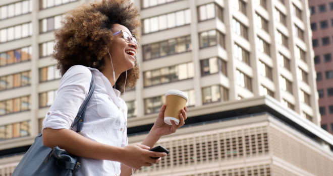 an african american woman smiles while listening to music, holding a coffee, and walking through a downtown area