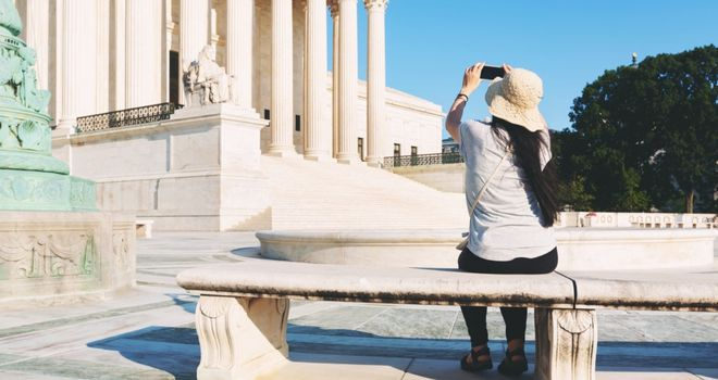 woman wearing a hat sits on a bench outside of the United States Supreme Court taking a picture with a small camera
