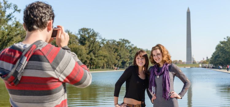 a man in a striped shirt take a picture of two women posing in front of the Washington Monument and reflecting pool in DC