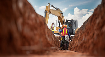 A construction crew walks toward a digger vehicle in a narrow trench